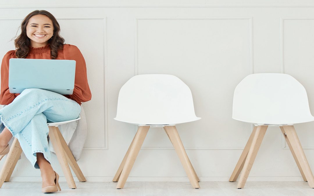 A cheerful woman sitting on a chair in a waiting room with a laptop on her lap, surrounded by empty white chairs against a minimalistic wall.